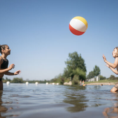 Zwei Kinder spielen mit einem bunten Wasserball im Wasser am Ufer.