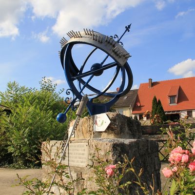 Sonnenuhr, die auf einem Steinsockel steht und von Blumen umrandet wird.
