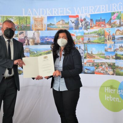 Landrat Thomas Eberth überreicht der Stellvertretenden Landrätin Christine Haupt-Kreutzer die Kommunale Dankurkunde des Bayerischen Innenministers. Beide tragen FFP2-Maske.
