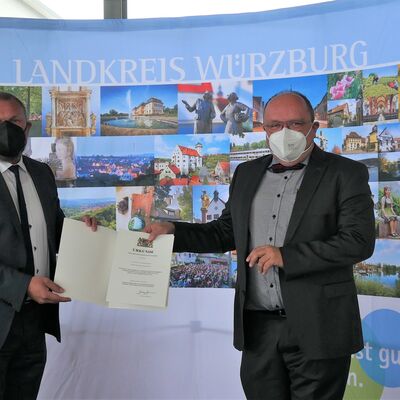 Landrat Thomas Eberth überreicht Prof. Dr. Alexander Schraml die Kommunale Dankurkunde des Bayerischen Innenministers. Beide tragen FFP2-Maske.