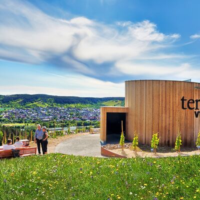 Rundes Holzgeb&auml;ude neben dem der Weinberg verl&auml;uft, in dem zwei Leute stehen.