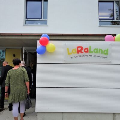 Mehrere Personen gehen durch eine Tür in ein Gebäude. An dem Haus hängen Luftballons und es ist die Aufschrift LaRaLand zu lesen.