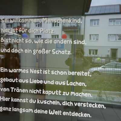 Ein Fenster auf der ein Gedicht aufgedruckt ist. Es lautet: Sei willkommen Menschen Kind, hier ist f&uuml;r dich Platz. Bist nicht so wie die andren sind, und doch ein gro&szlig;er Schatz. Ein warmes Nest ist schon bereitet, gebaut aus Liebe und aus Lachen, von Tr&auml;nen nicht kaputt zu machen. Hier kannst du kuscheln, dich verstecken, ganz langsam deine Welt entdecken.