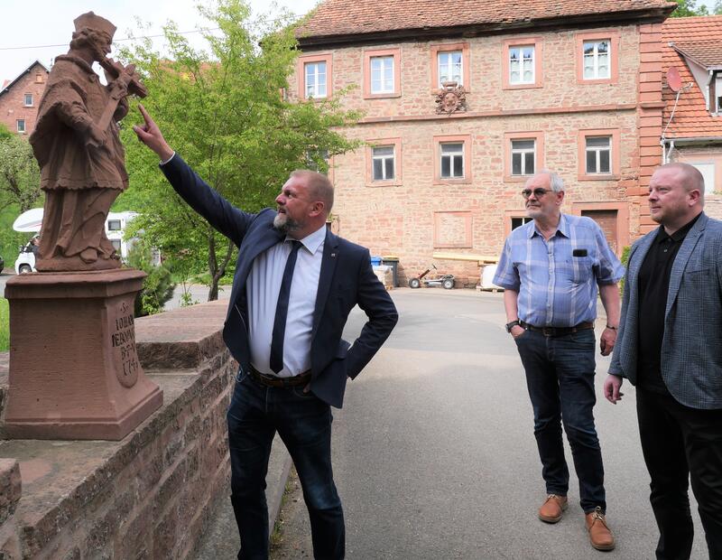 Bild vergrößern: Holzkirchen ist stolz auf seine Baudenkmäler. Landrat Thomas Eberth (links) bewundert die Nepomuk-Statue aus rotem Buntsandstein, dem prägenden Baustoff im Ort. Im Hintergrund das Gebäude der ehemaligen Klostermühle.